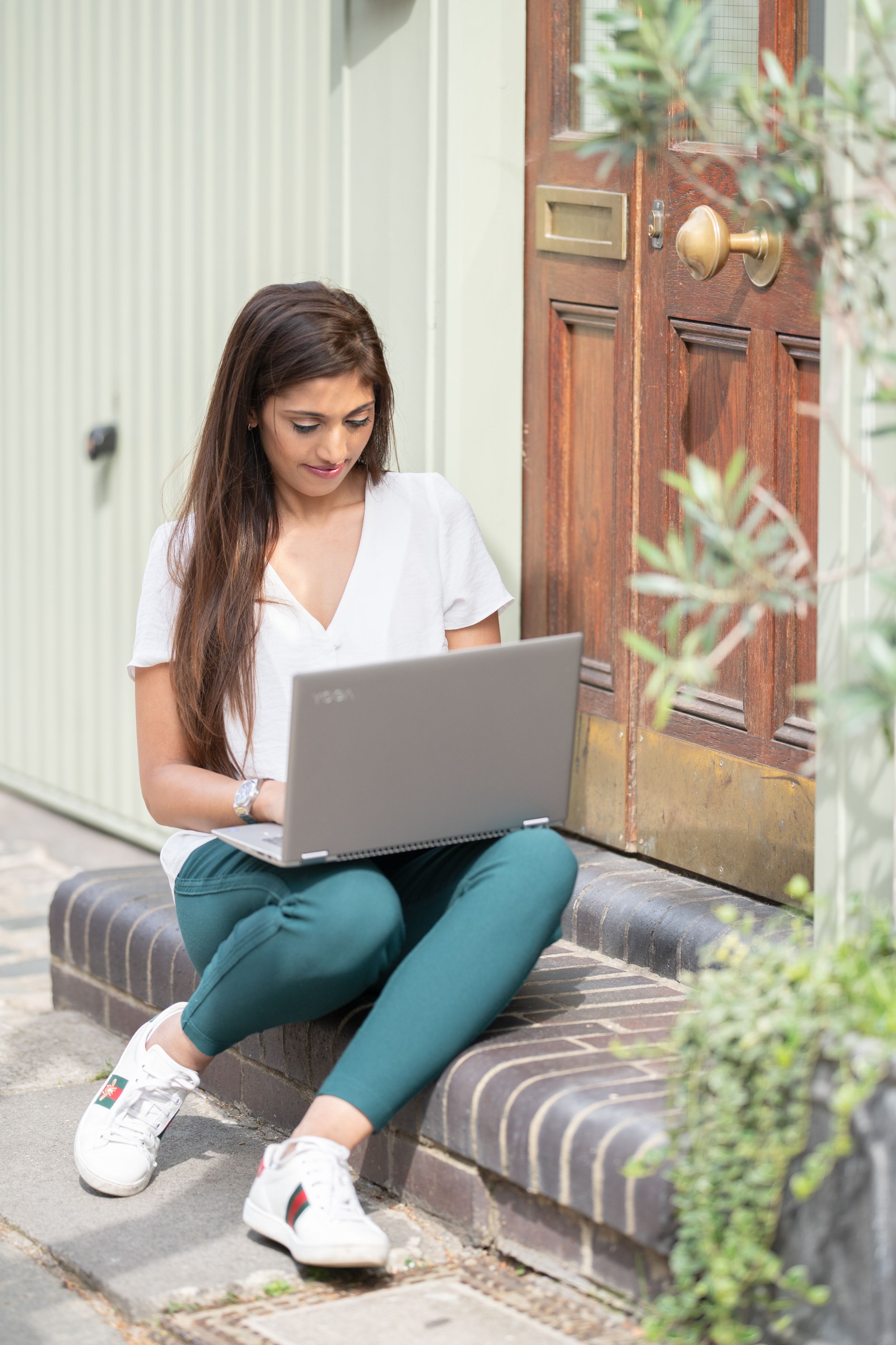 Person in white shirt and green pants working on laptop while sitting on outdoor steps near plants and wooden doors.