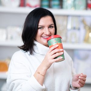 Person in white sweater holds and examines a small red cosmetic product container in bright indoor setting.