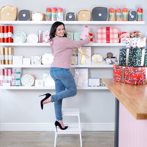 A person in pink sweater and jeans stands on a step stool reaching for items on white shelves filled with colorful containers.