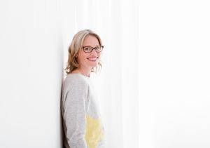 A smiling person wearing glasses and a gray sweater leans against a white wall.