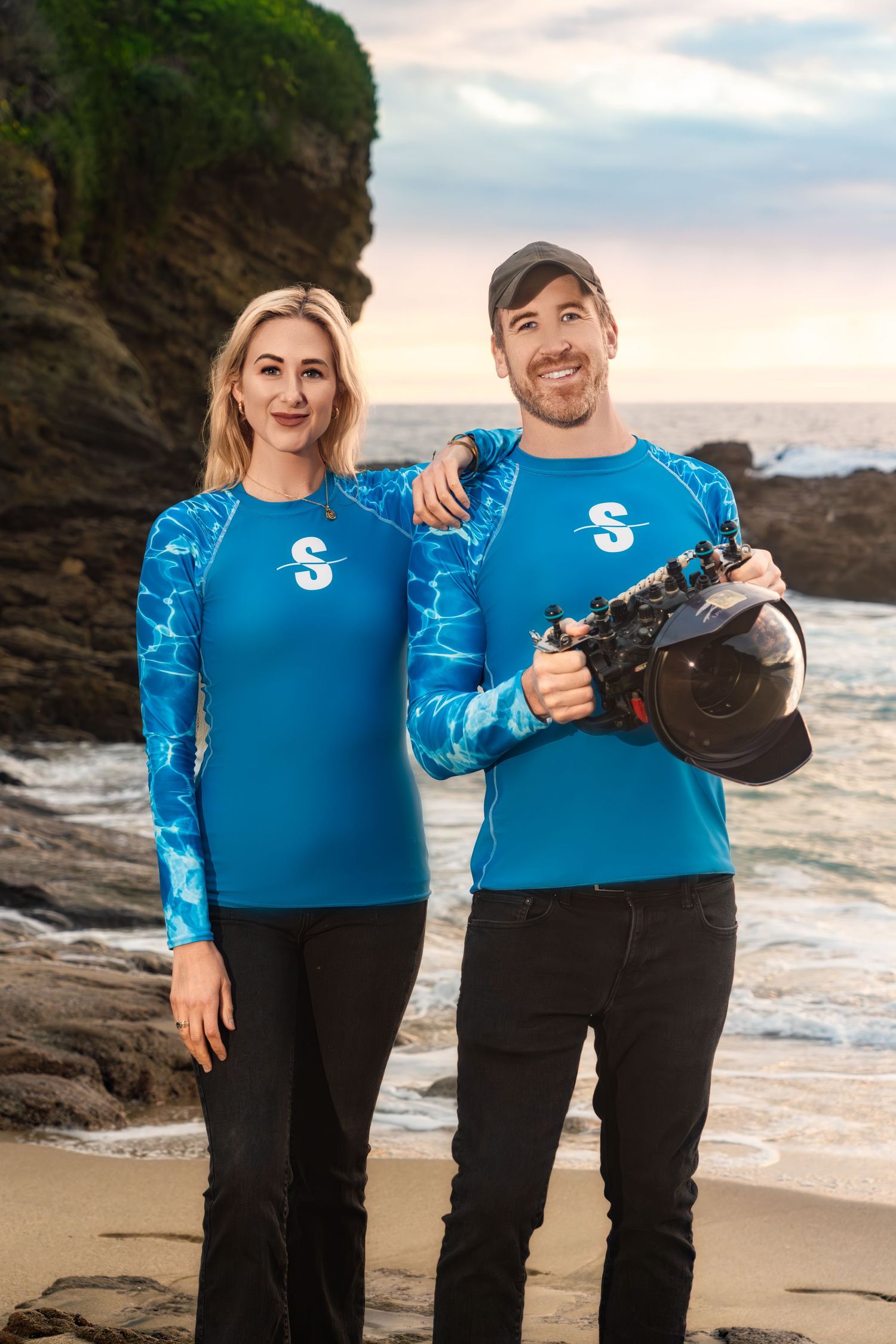 A couple wearing matching blue rash guards poses together on a beach at sunset near rocky cliffs.