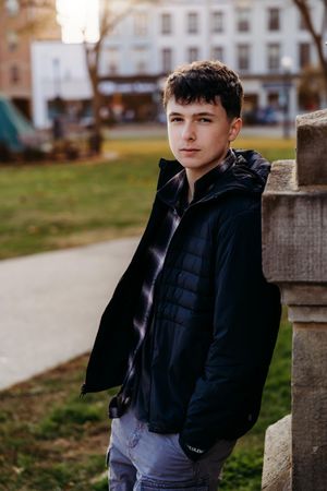 Portrait of someone wearing a navy blue puffer jacket and jeans leaning against a stone pillar in an outdoor setting.