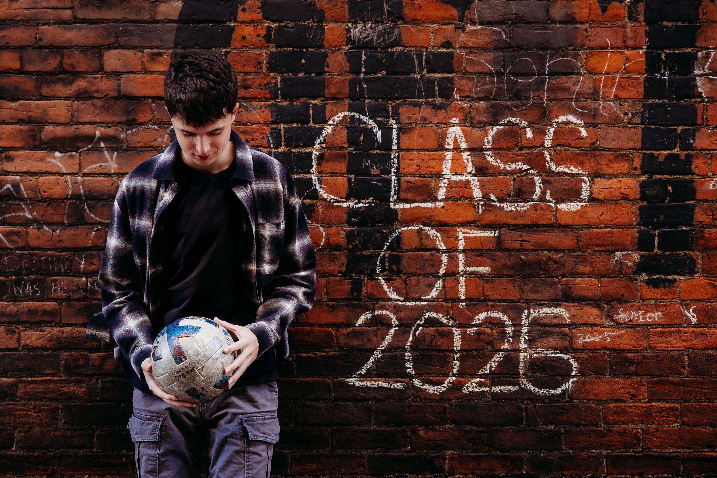 Student holding a soccer ball stands against brick wall with 'Class of 2026' graffiti in moody lighting.