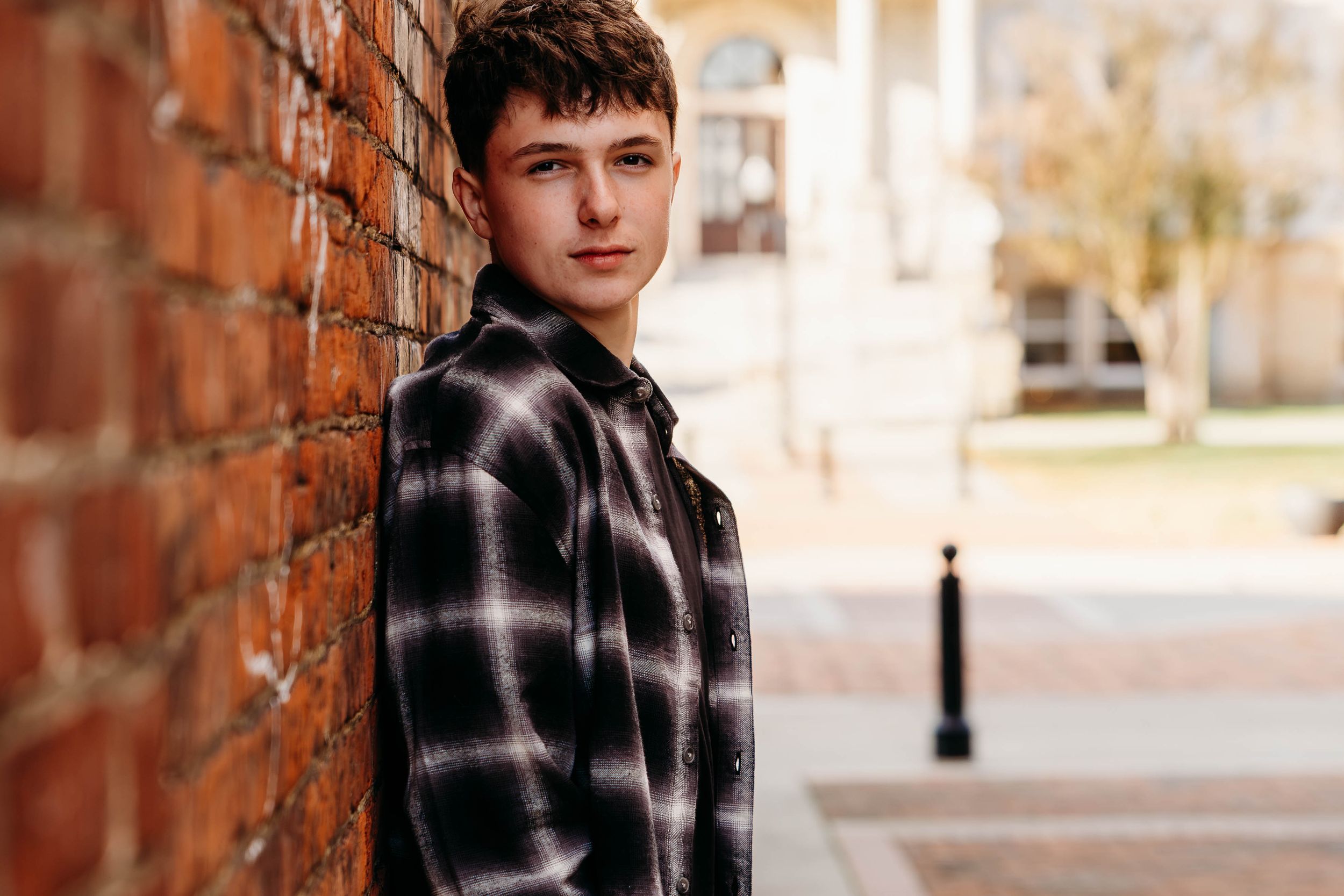 A person in a black and white plaid flannel shirt poses against a brick wall in warm autumn lighting.