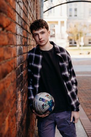 Young person in plaid flannel and dark clothes poses with soccer ball against brick wall in outdoor setting.