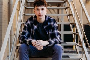 A person in a dark blue plaid shirt sits casually on wooden steps in an industrial or warehouse setting.
