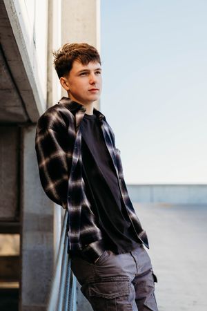 A person in a dark jacket and plaid shirt poses thoughtfully against a light-colored wall in a minimalist setting.