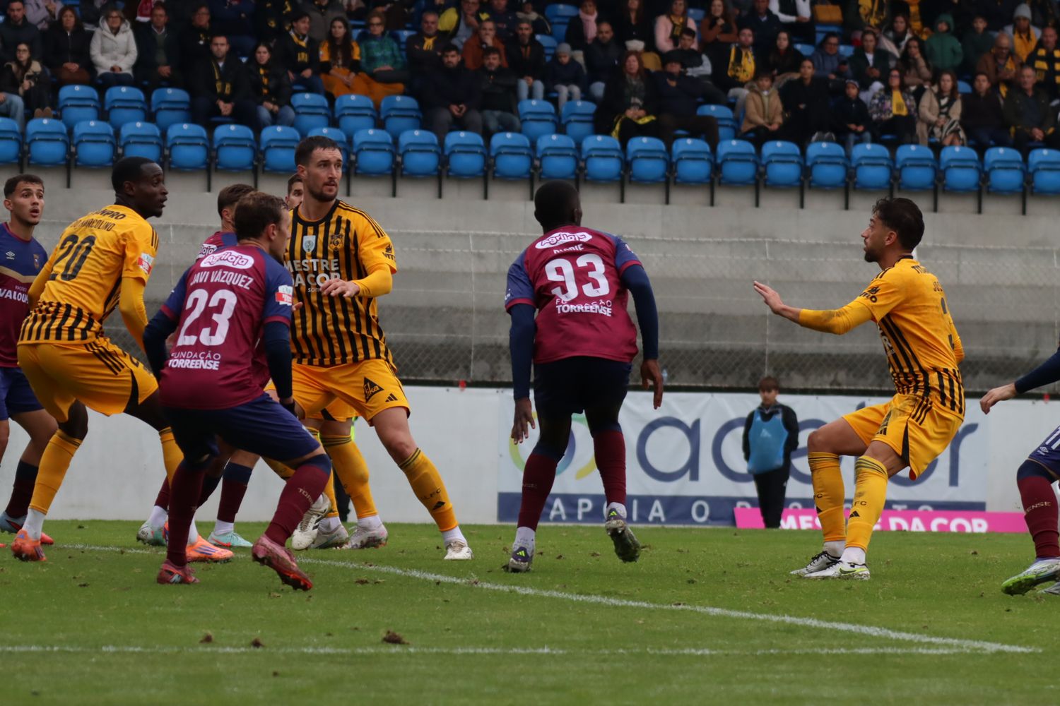 Soccer match action between teams in yellow and purple uniforms with stadium seating visible.