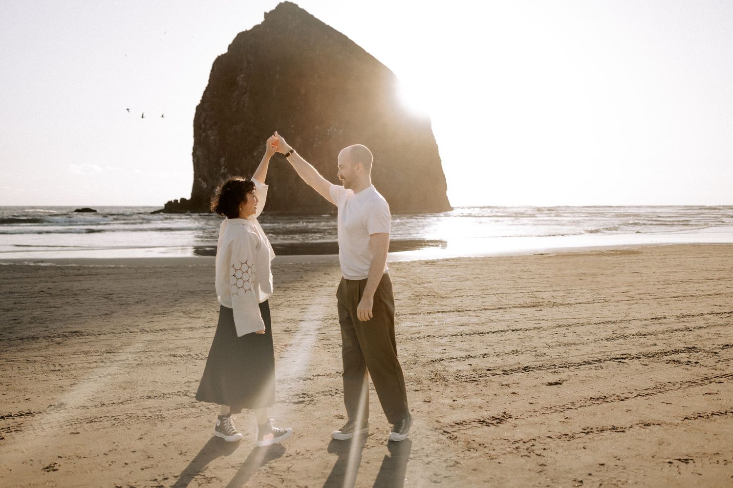 Romantic couple walking along a sunlit beach at sunset with ocean waves and cliffs in the background.