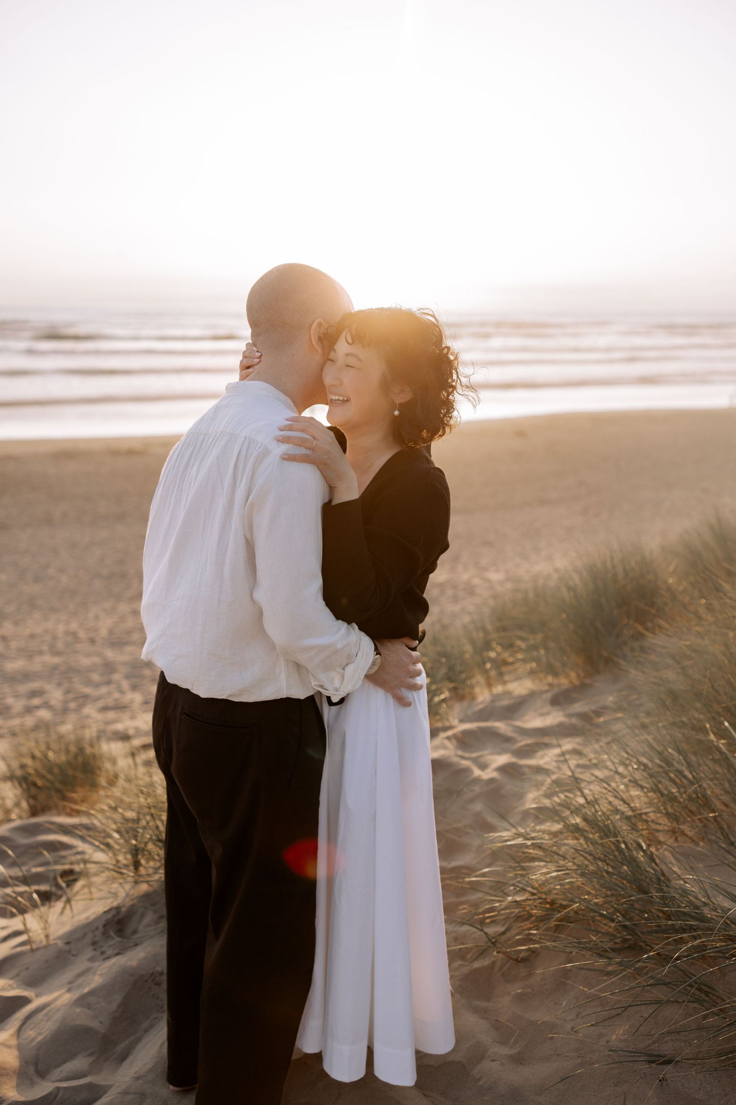 A romantic couple embraces on a beach at sunset, with the woman in black and white flowing dress against golden light.