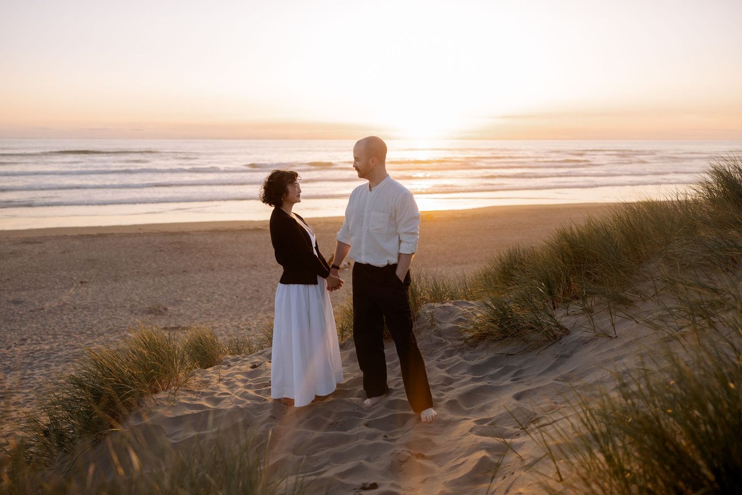 Romantic sunset beach photoshoot with couple standing against golden sky and sand dunes.
