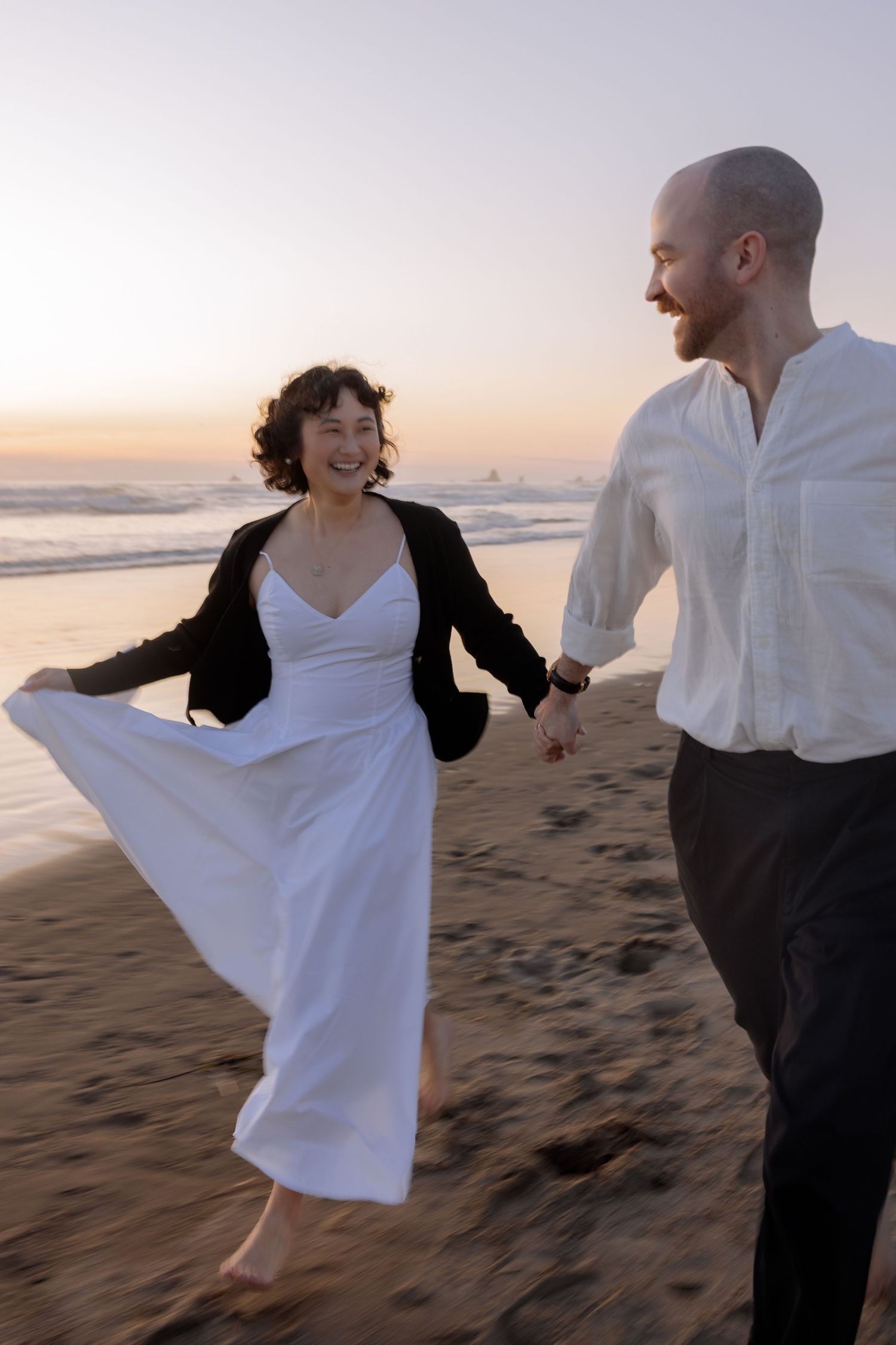 A couple in black and white outfits dance joyfully together on a beach at sunset, creating romantic silhouettes.