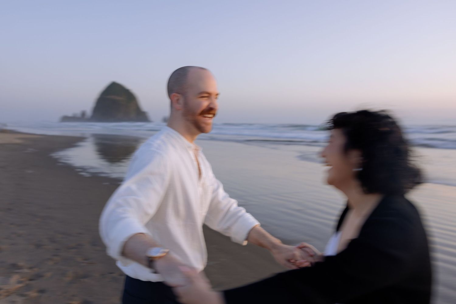 A couple dances together on the beach with Haystack Rock visible in the background at dusk.