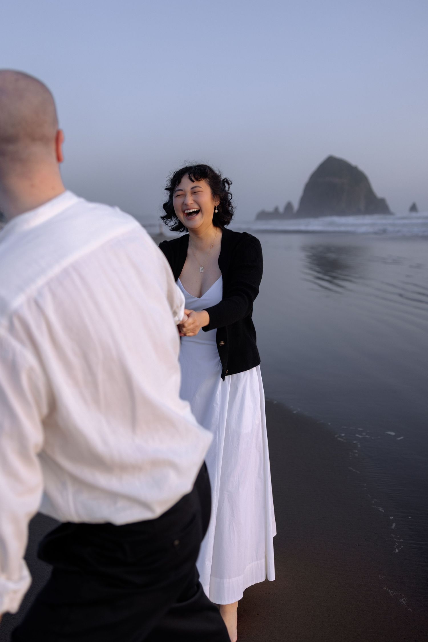 A couple embraces on a misty beach with dramatic coastal rock formations in the background.