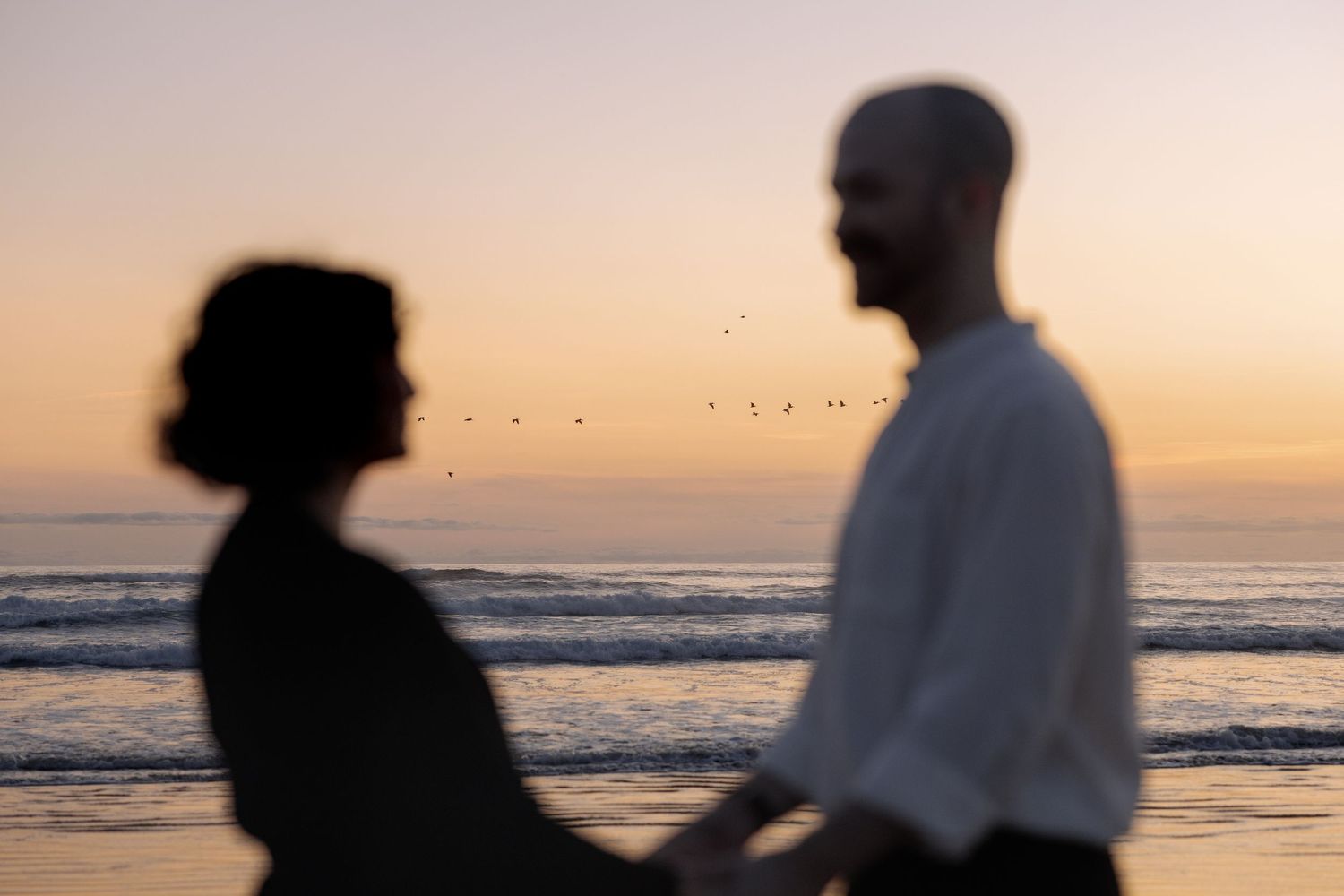 Silhouettes of two people standing close together on a beach at sunset with ocean waves in the background.