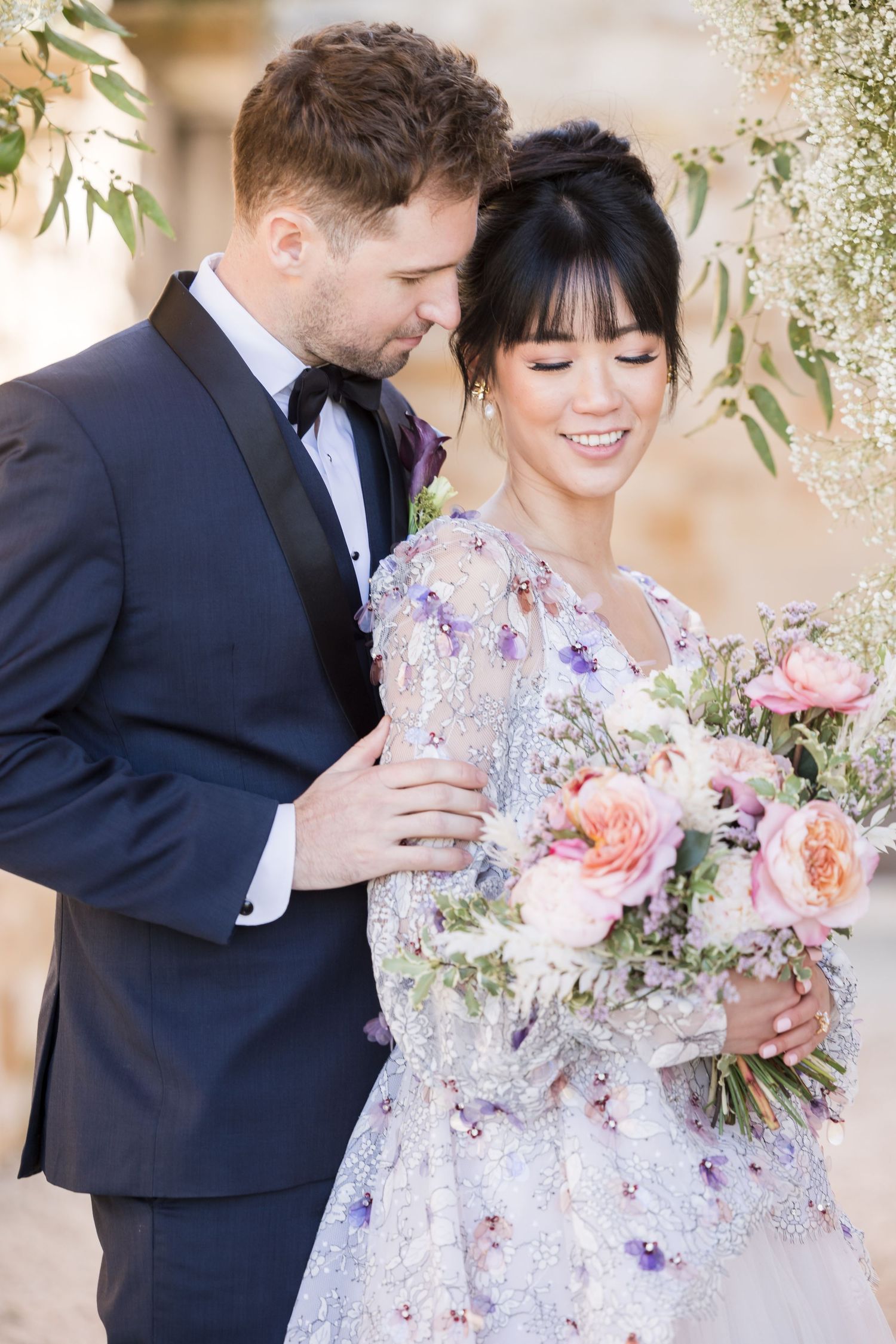 A couple in white wedding attire walks together along a tree-lined path in a serene park setting.