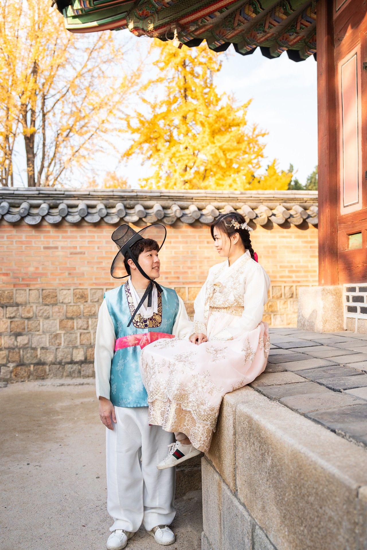 Two people in traditional Korean hanbok outfits interact at a historic stone wall during autumn with golden foliage.