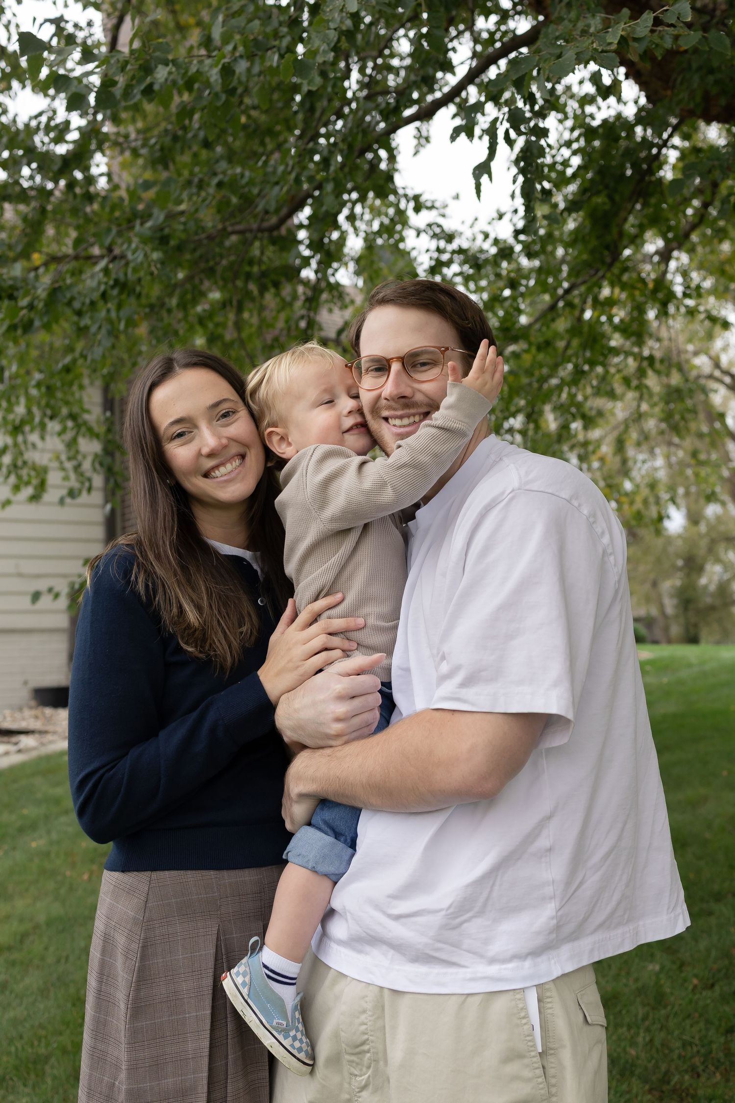 Sioux City, Iowa photographer. A smiling young family with a toddler posing together outdoors under a green tree.
