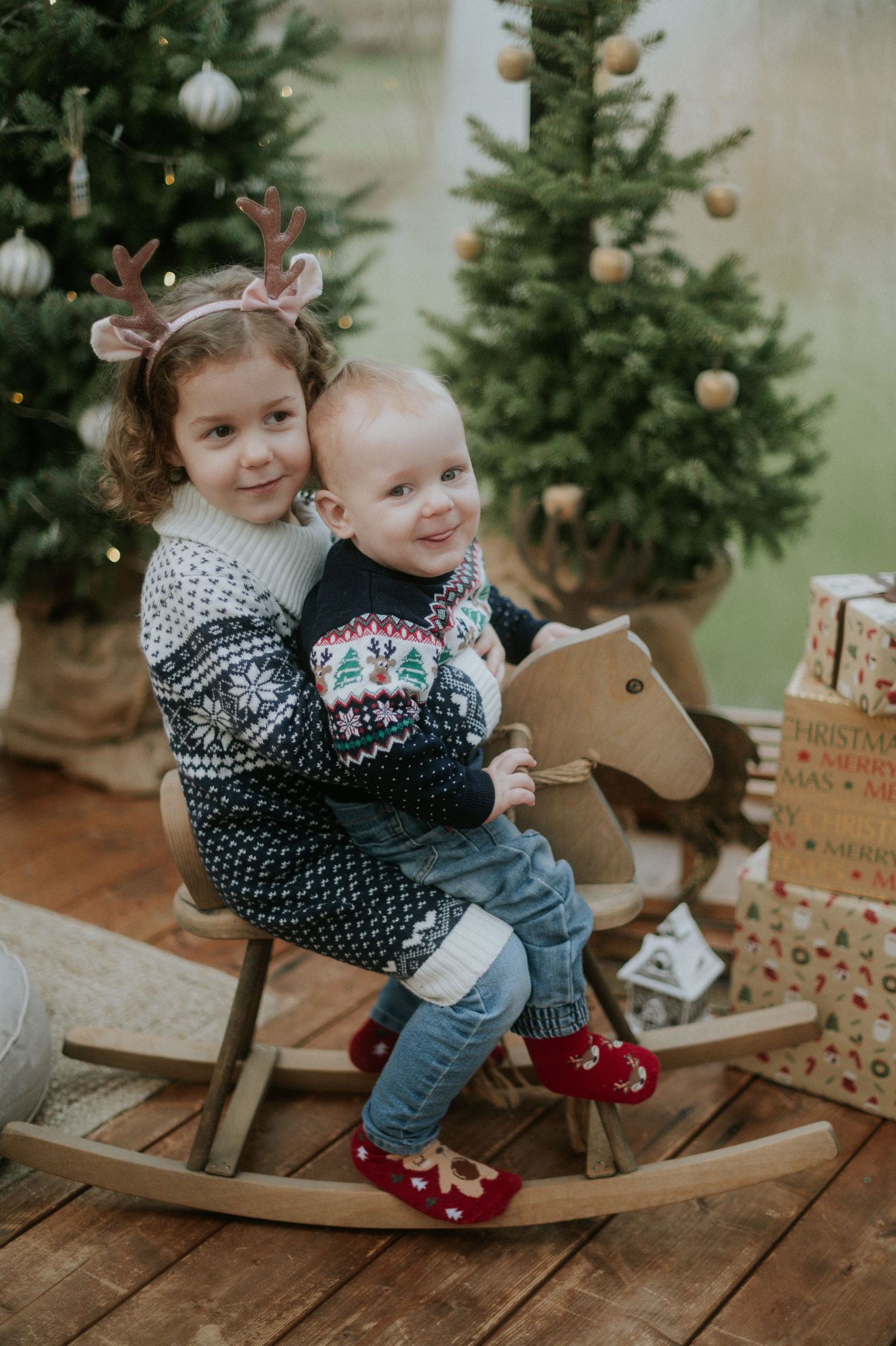 Two children wearing winter sweaters play together on a wooden rocking horse by a decorated Christmas tree.