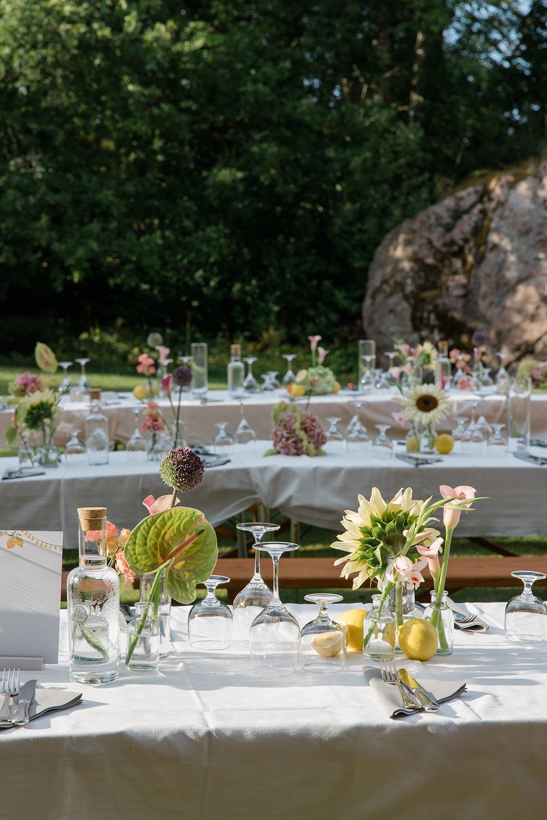 Elegant outdoor wedding reception tables set with white linens, floral centerpieces and glassware in a garden setting.