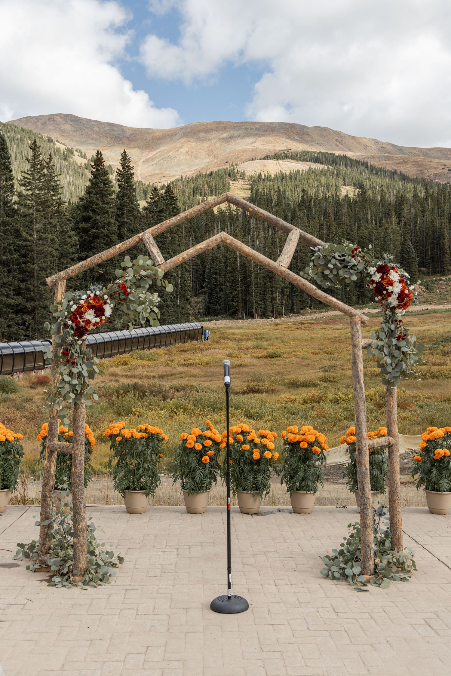 A rustic wooden wedding arch decorated with red flowers and greenery stands against a mountain landscape backdrop.