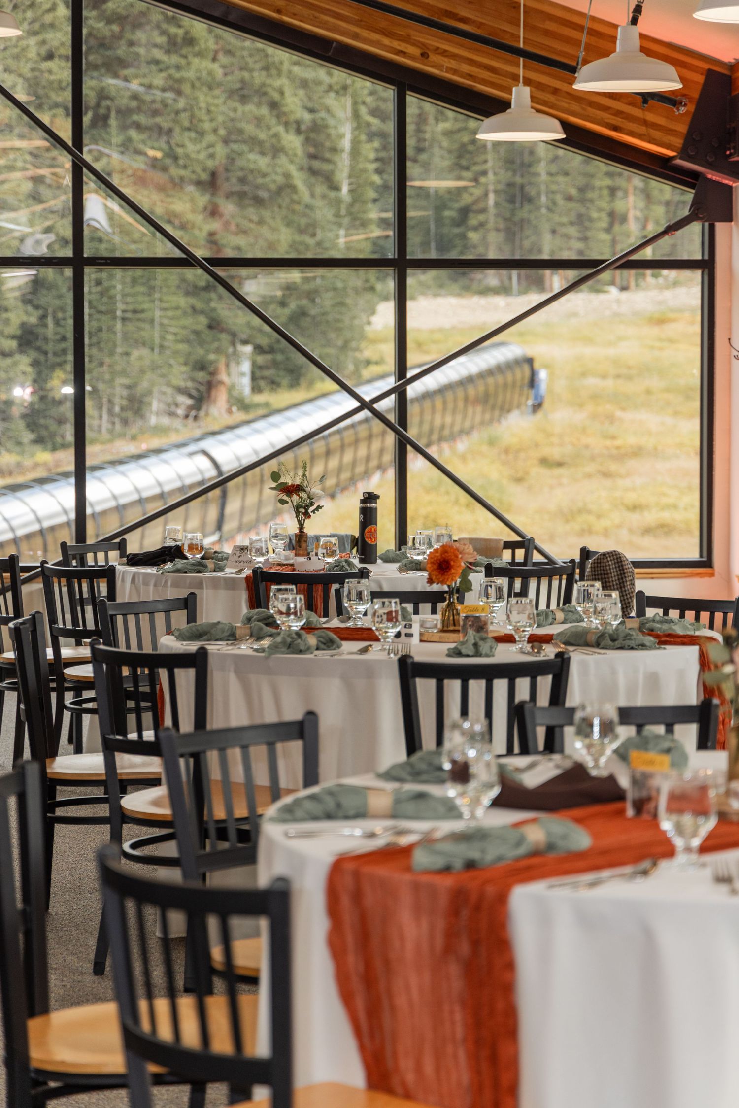A rustic mountain lodge dining room with white tablecloths and black chairs overlooking scenic mountain views through windows.