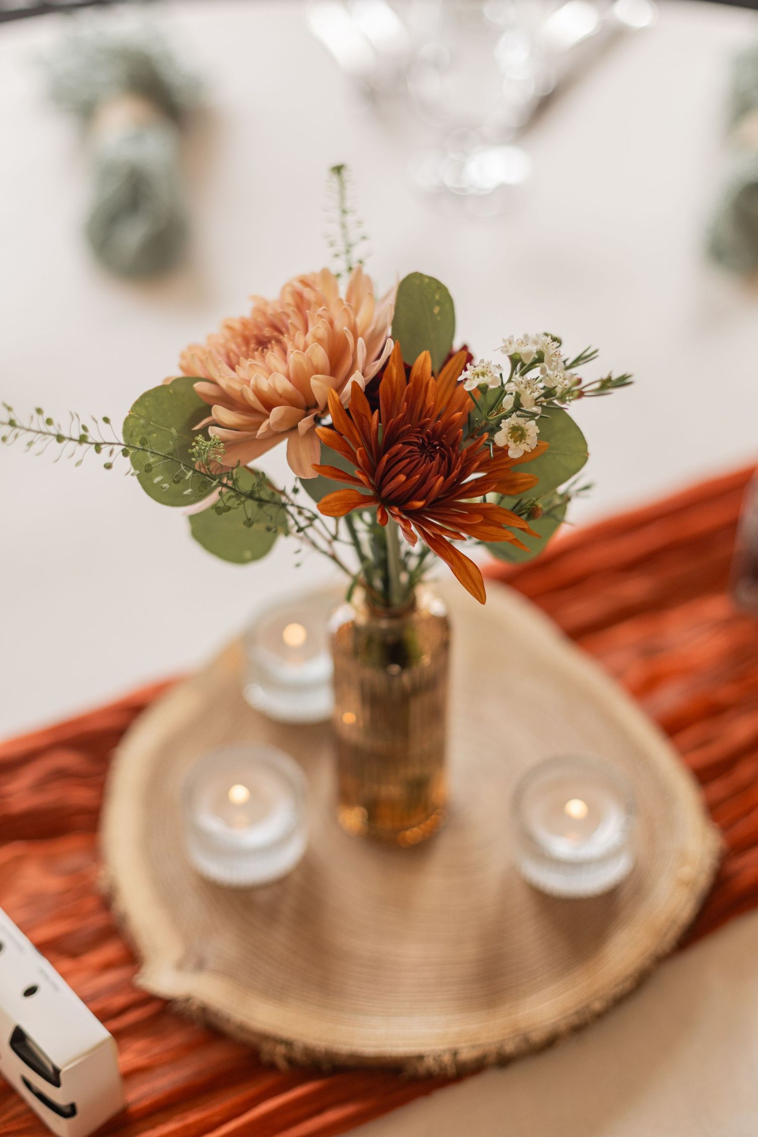Orange chrysanthemum flowers in small glass vases on wooden slices with glowing tea lights create an elegant table setting.