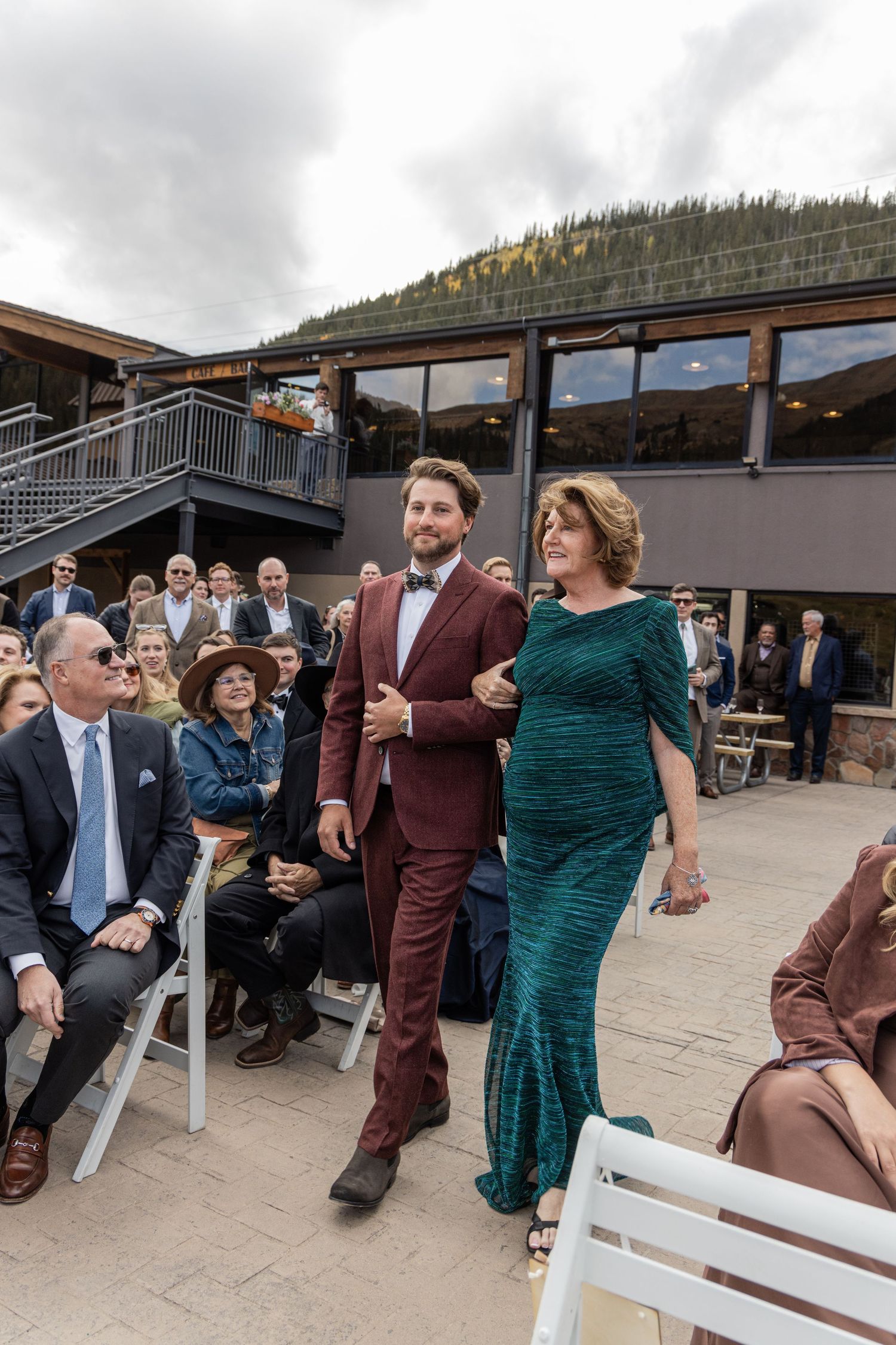 Wedding attendees gather at an outdoor mountain venue as a couple walks down the aisle in burgundy suit and emerald dress.