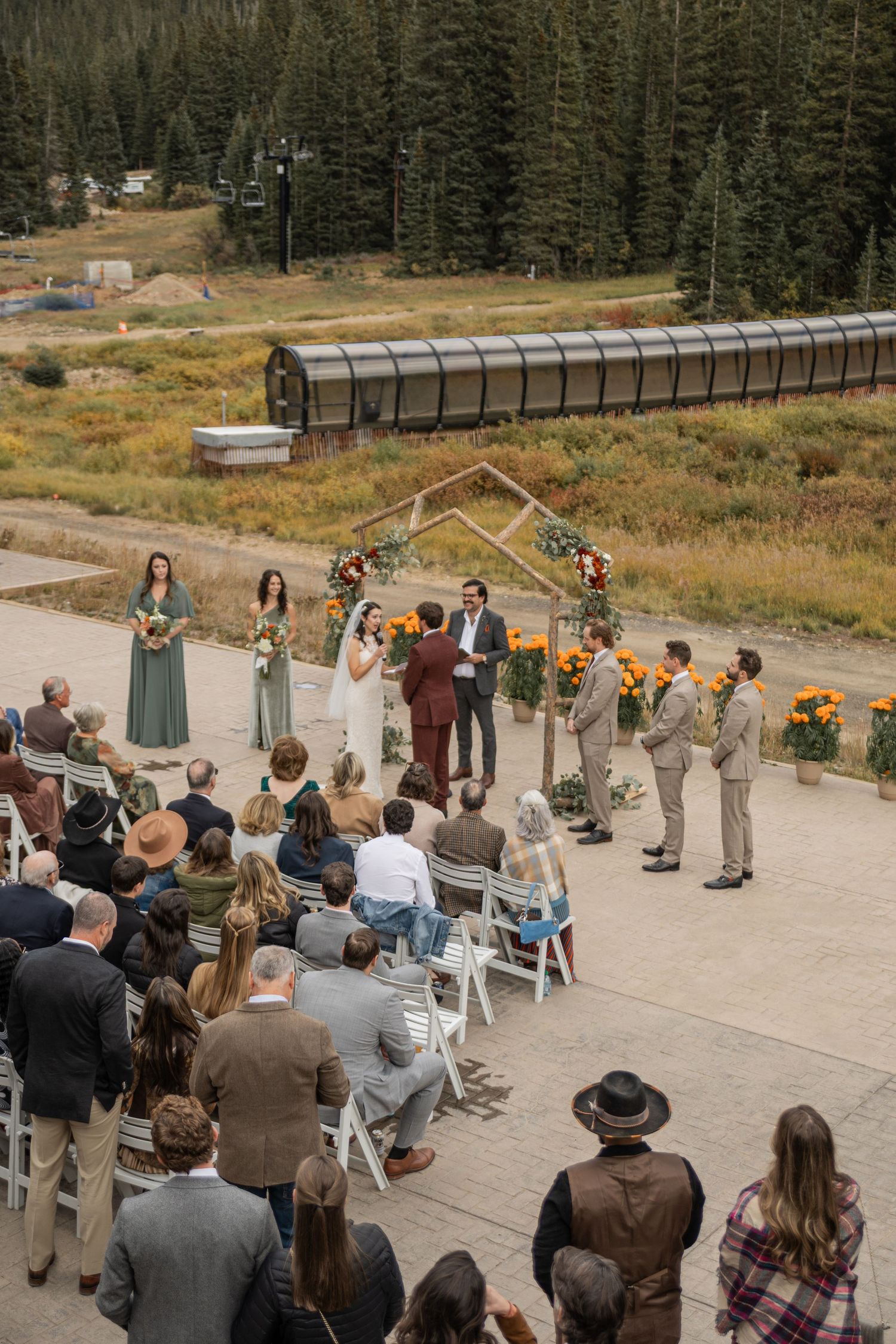 A small outdoor wedding ceremony setup with guests seated in white chairs surrounded by mountain scenery and evergreen trees.