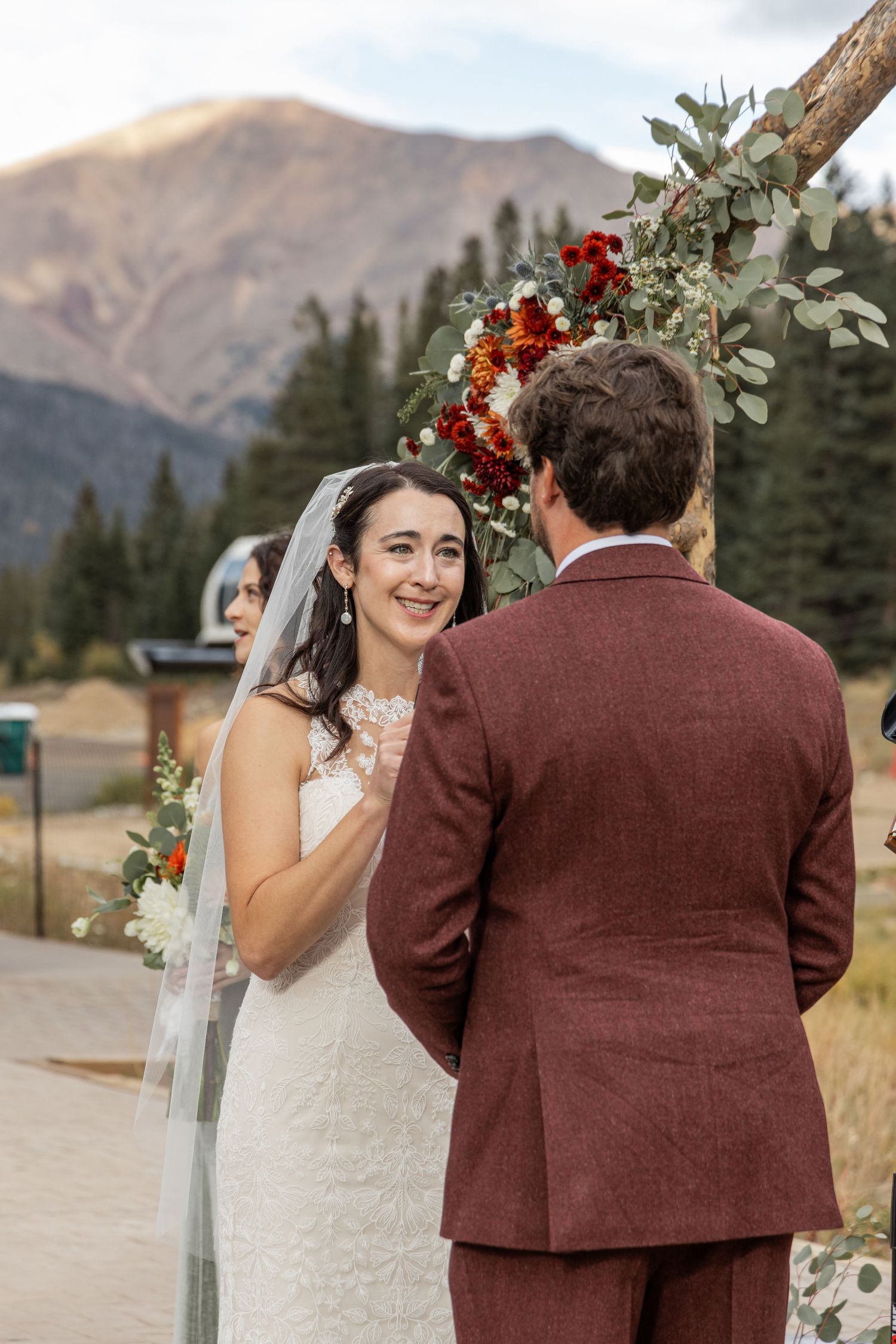 A wedding ceremony takes place outdoors in the mountains with burgundy and floral decor details.