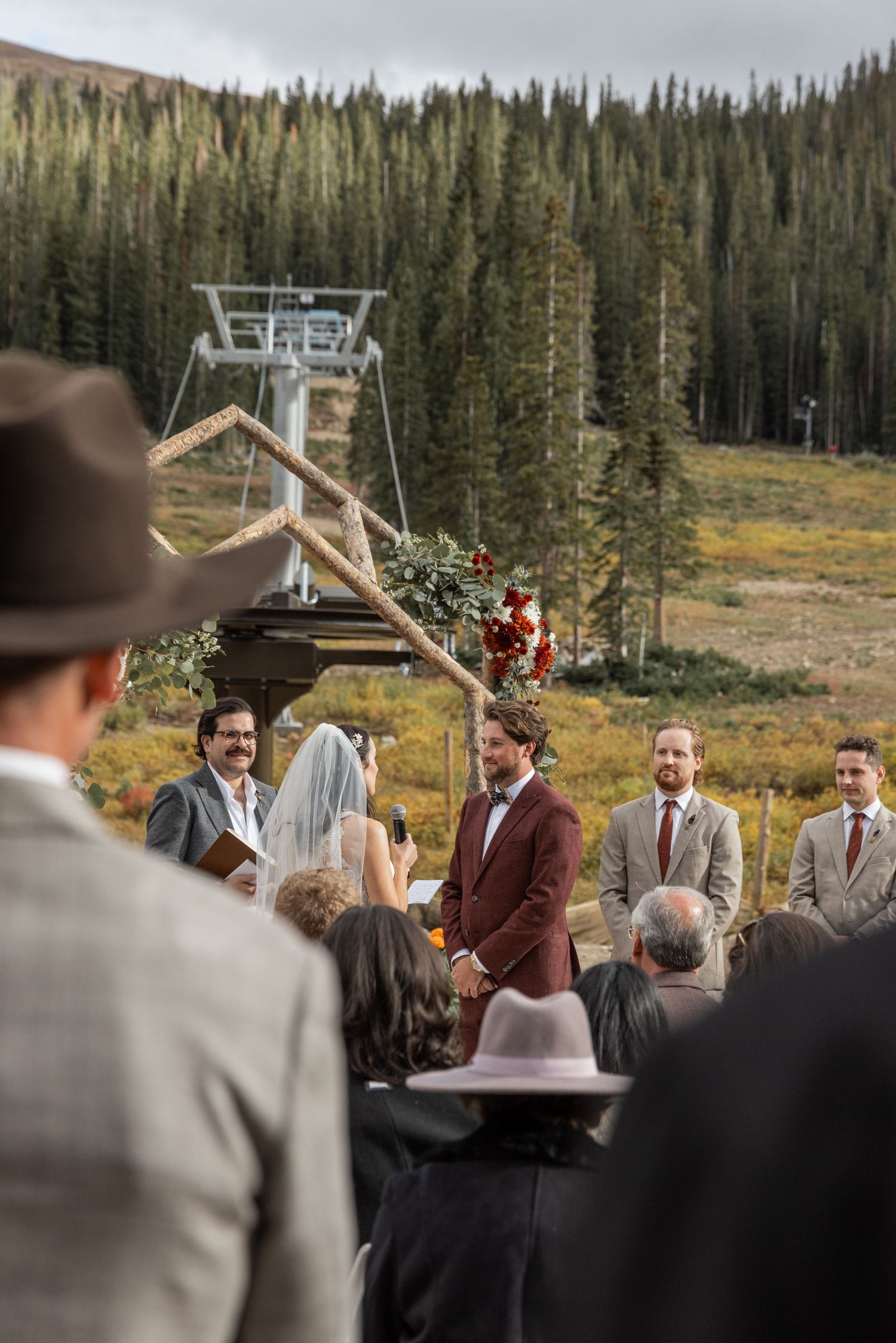 A wedding ceremony takes place outdoors at a ski resort surrounded by evergreen trees and mountains in the background.