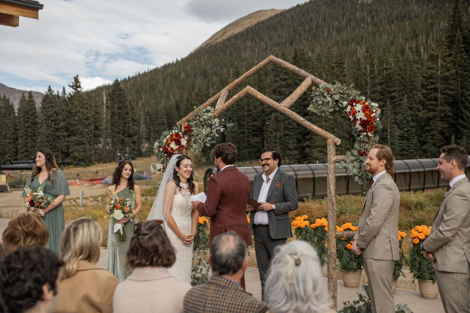 Wedding ceremony guests gather outdoors at a mountain venue with pine trees and rustic wooden structures in the background.