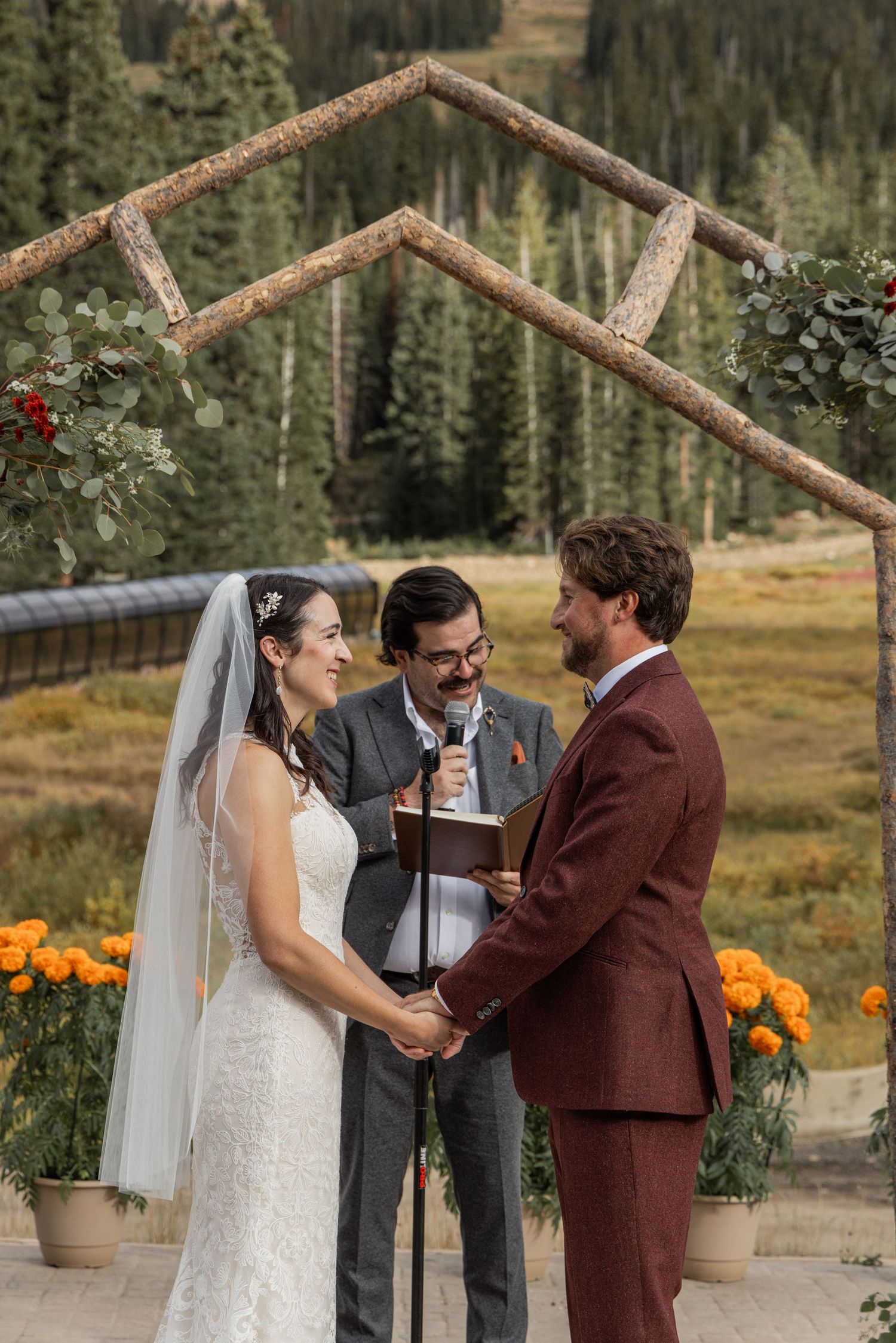 A couple exchanges vows during an outdoor mountain wedding ceremony with orange flowers and evergreen trees in background.
