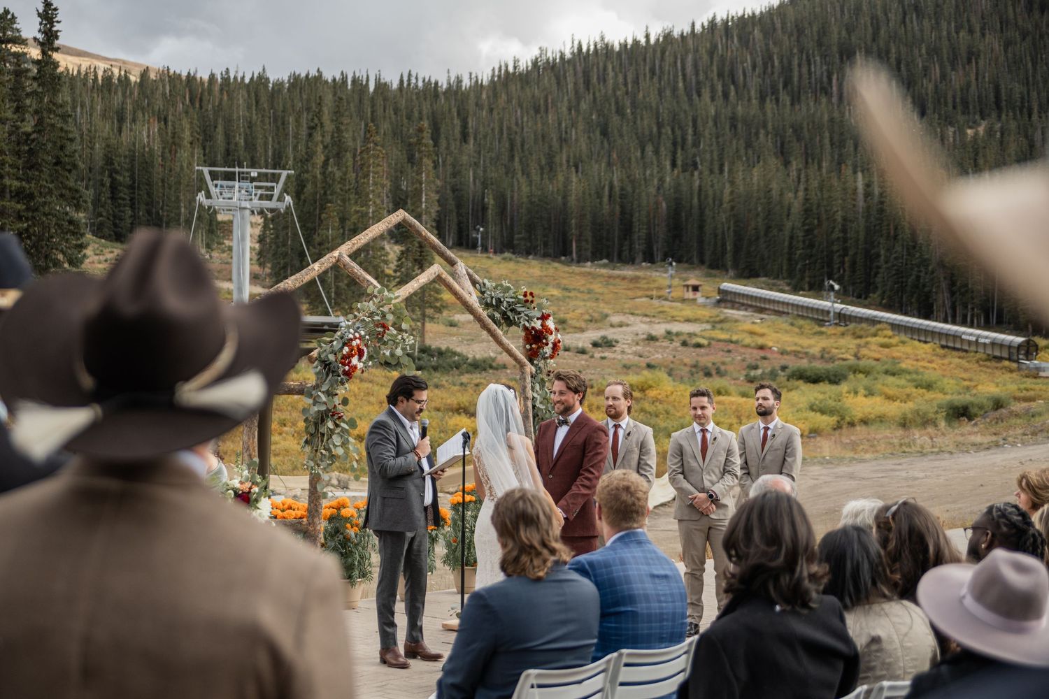 A scenic mountain wedding ceremony takes place outdoors with chairs set up in front of snow-capped peaks and evergreen trees.