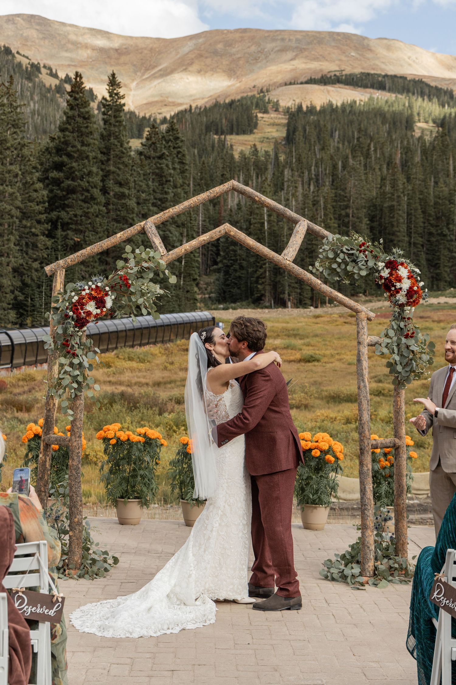 A romantic mountain wedding ceremony takes place under a rustic wooden arch with fall flowers in Colorado.