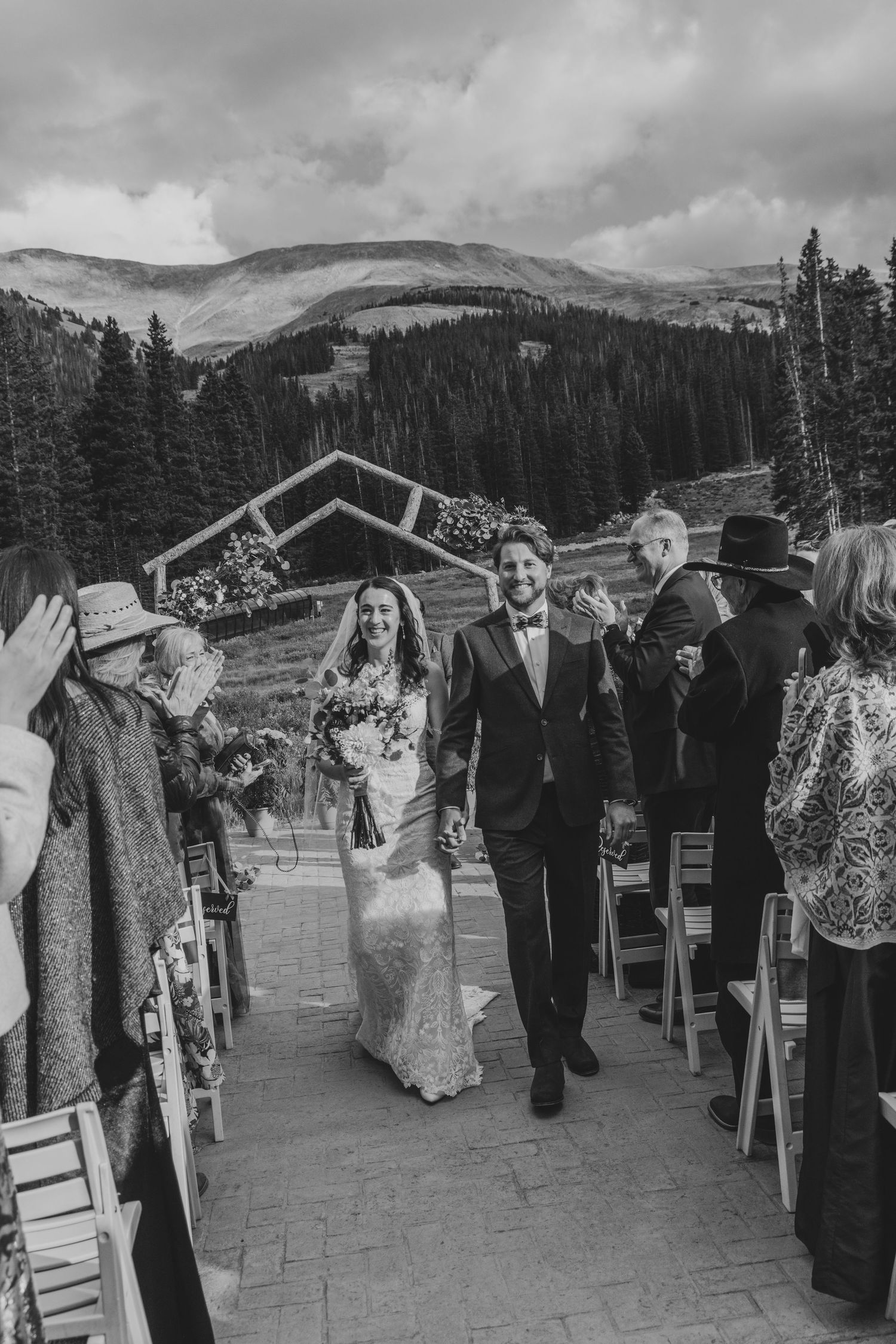 Wedding guests walk in a line along a mountain path with scenic snow-capped peaks and evergreen forests in the background.