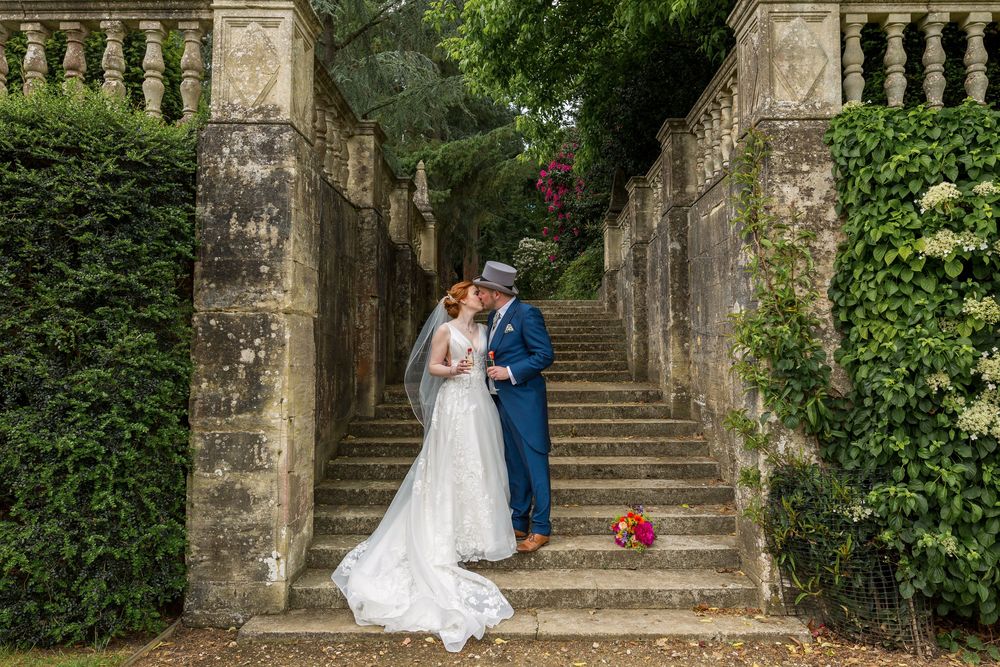 A couple in formal wedding attire embraces on a grand stone staircase surrounded by lush greenery and architecture at Tylney Hall