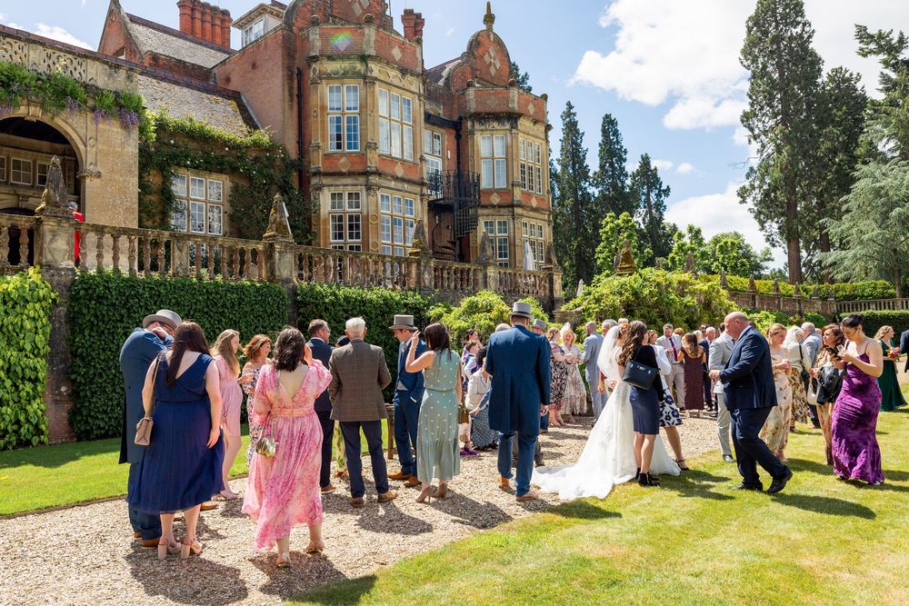 Wedding guests gather on a sunny lawn outside a grand Victorian mansion with ivy-covered walls during a summer celebration at Tylney Hall