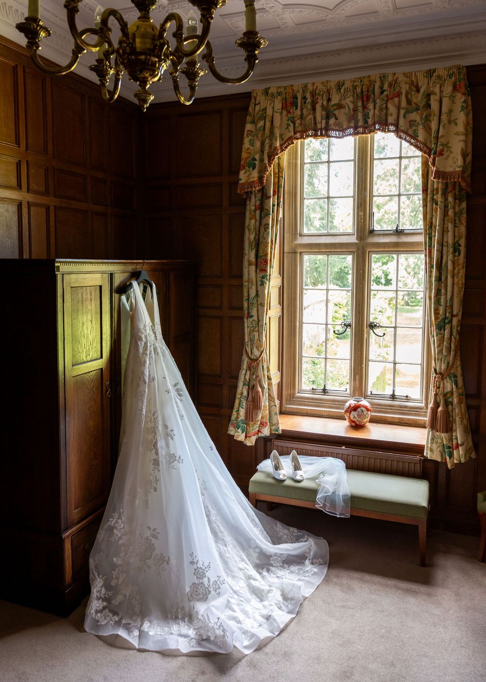 A white wedding dress with lace details and long train displayed in a wood-paneled room with chandelier and floral curtains at Tylney Hall