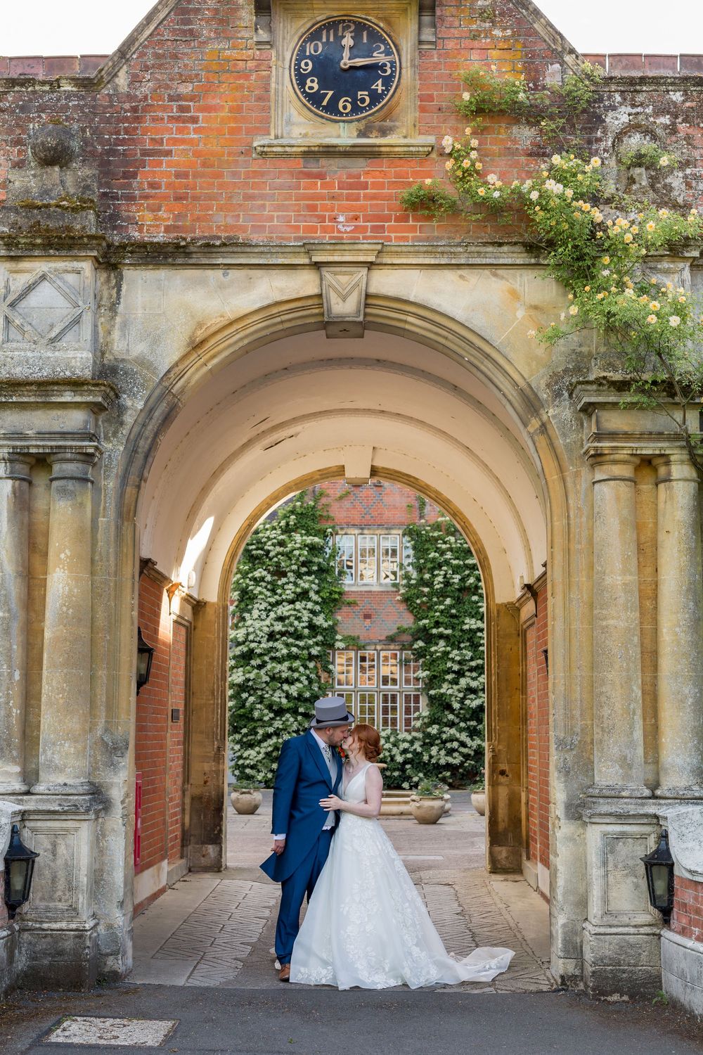 A couple shares a romantic moment under stone archways at a historic brick building with a clock tower at Tylney Hall