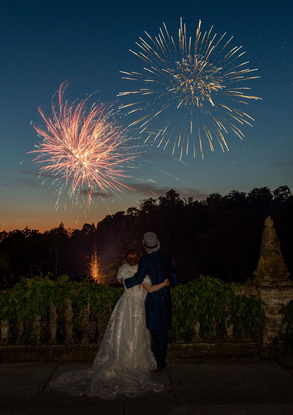 A couple watches fireworks burst in vibrant colors across the twilight sky over the grounds of Tylney Hall