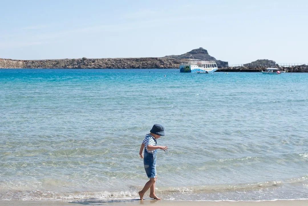 A child walking along the shoreline in clear shallow water with rocky islands in the distance.