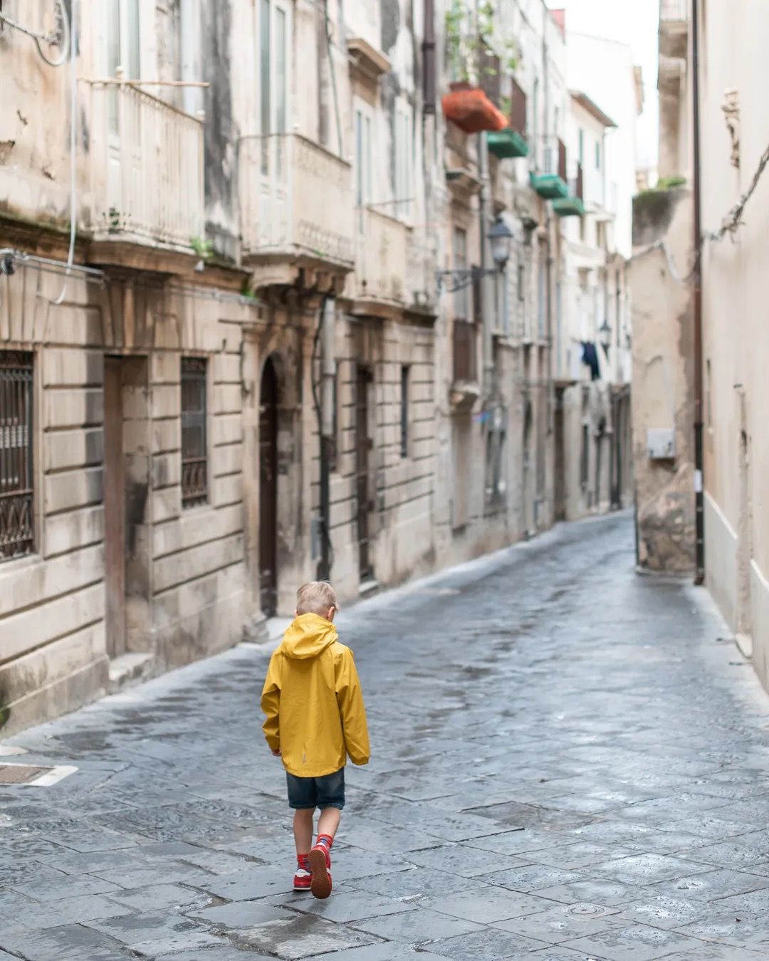 Woman in a yellow coat walking alone down a narrow historic street lined with tall stone buildings.