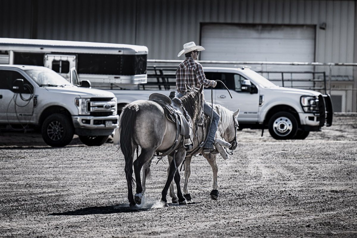 Monochrome image of person on horseback near parked vehicles.