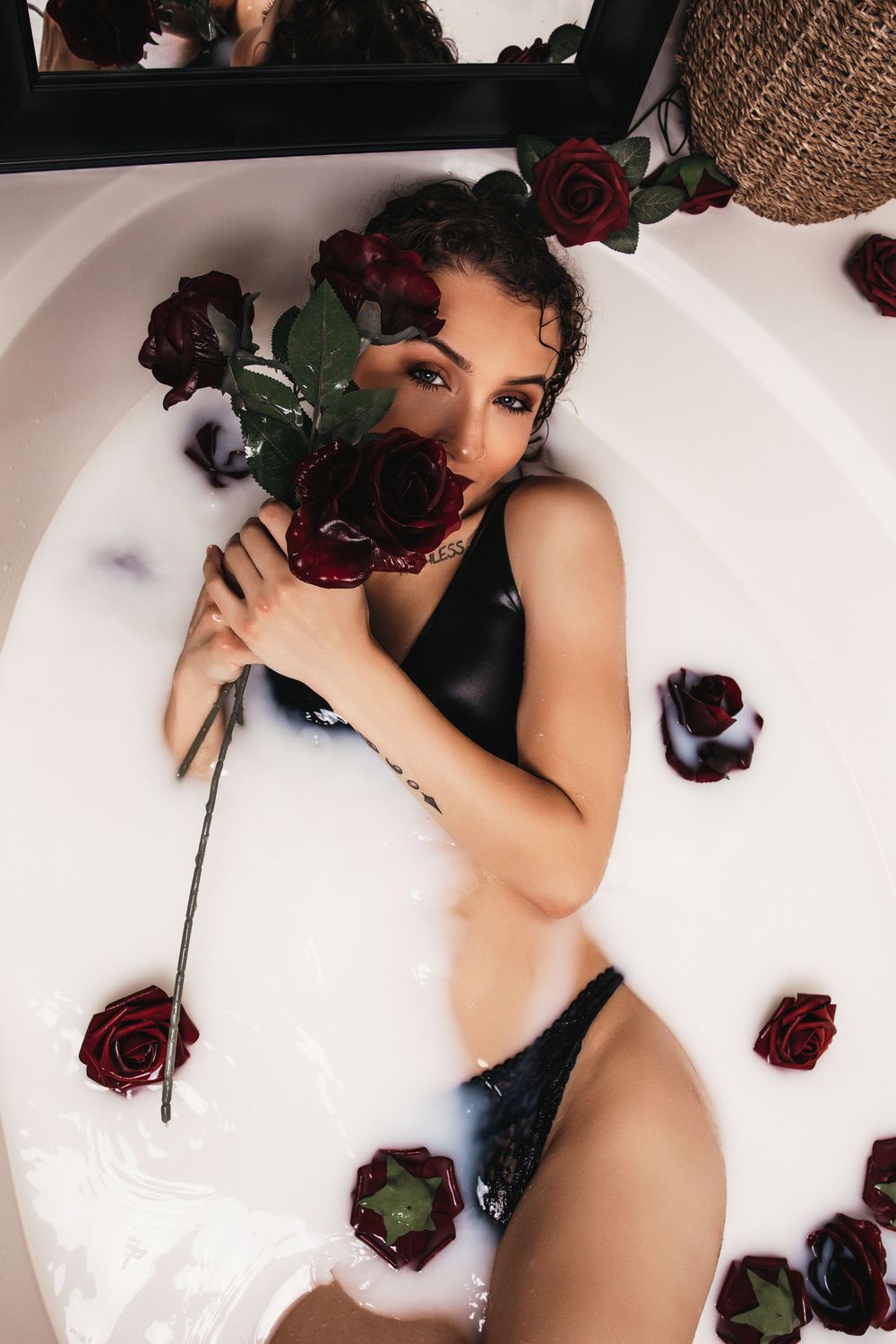 A milk bath photoshoot featuring dark red roses floating around a figure in white water.