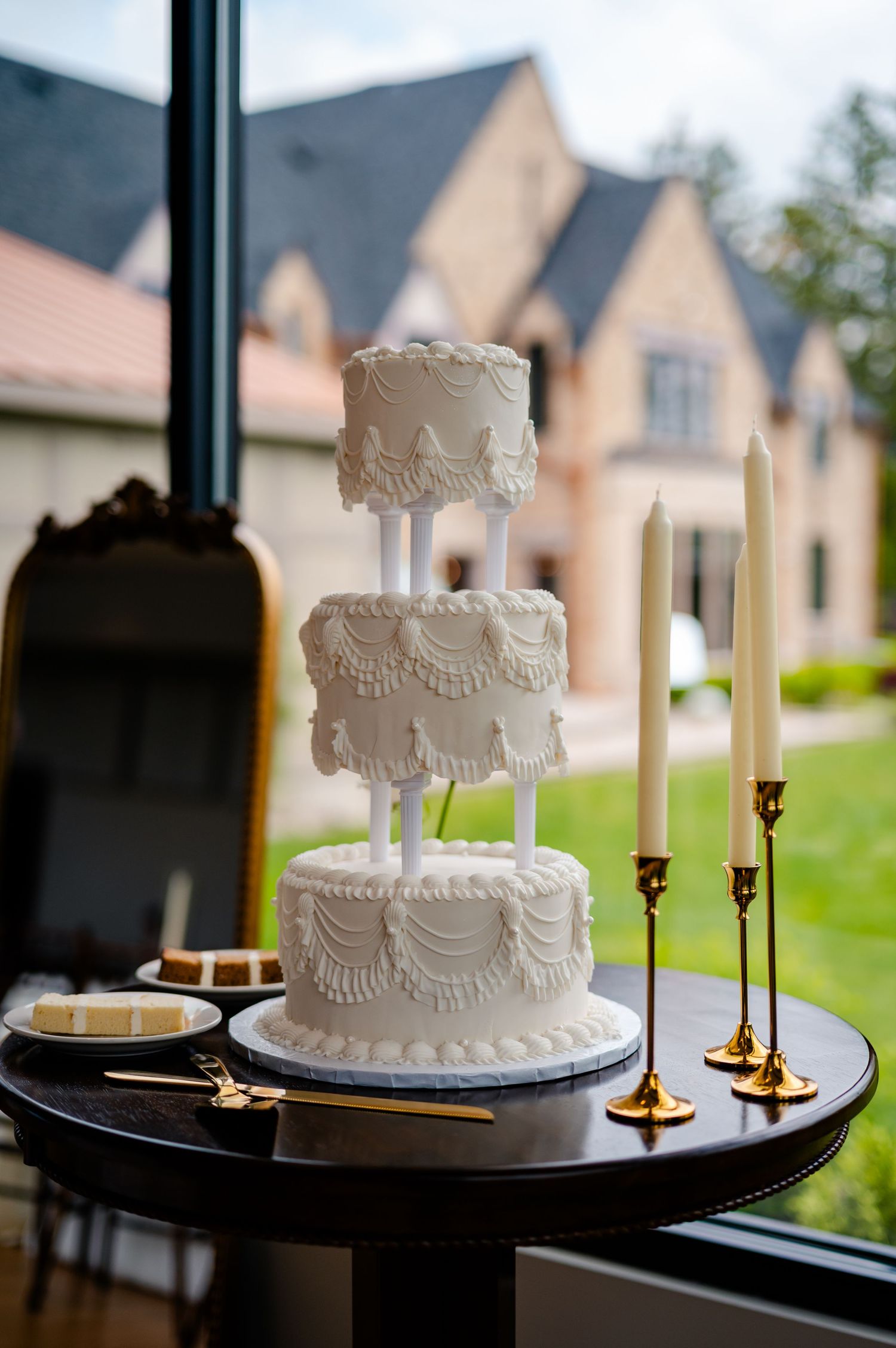 A white wedding cake with delicate piping sits elegantly on a dark tray with brass candlesticks in a window setting.