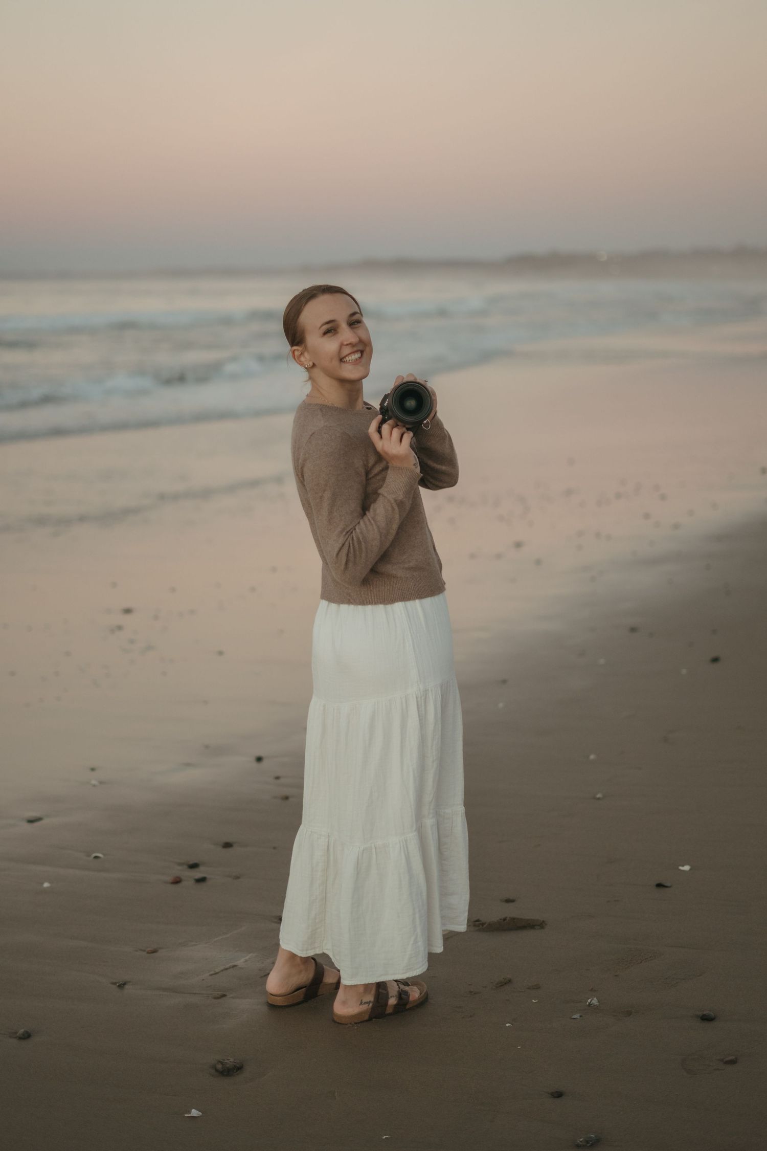 Person in beige sweater and white dress dances joyfully on a misty beach at sunset.