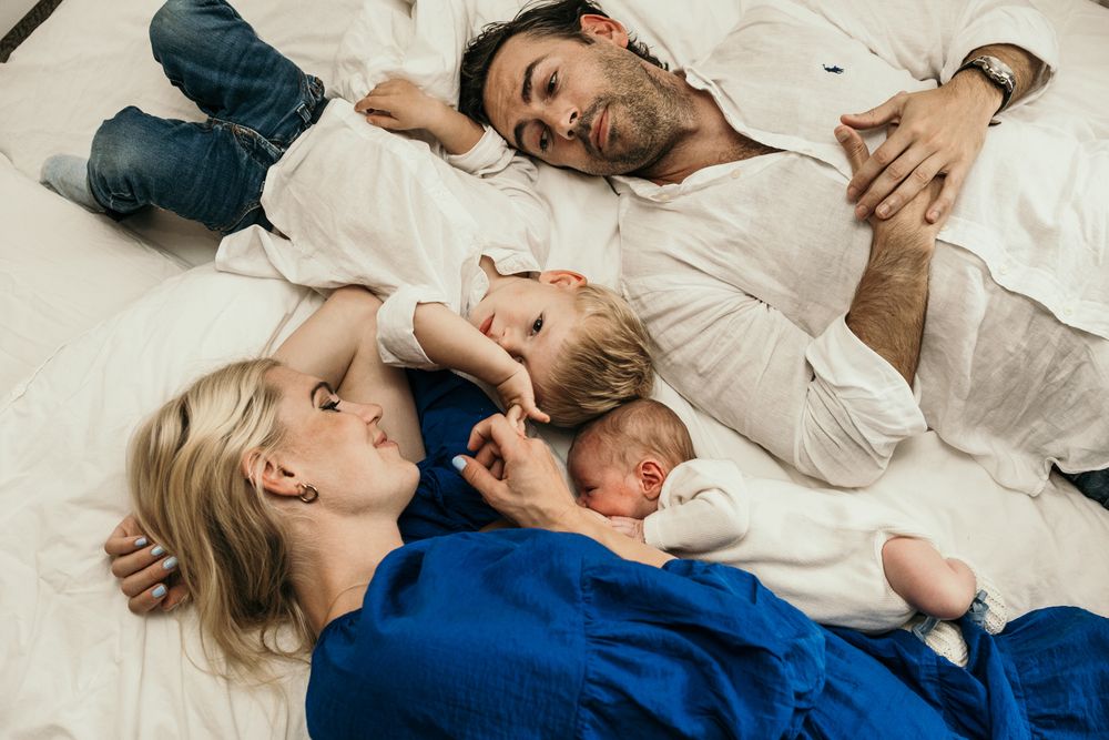 A family rests together on white bedding, with one person in a blue outfit and another in white clothing.