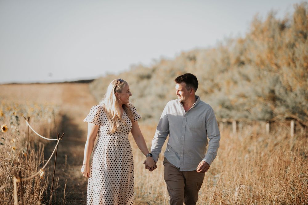 Smiling couple holding hands walking through golden meadow fields on a sunny day.