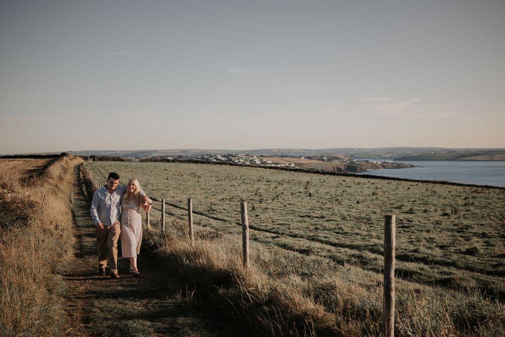 A couple walks along a coastal path beside a wooden fence with expansive green fields and ocean views.